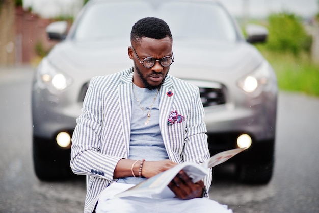 rich-stylish-african-american-man-blazer-white-pants-eyeglasses-with-magazine-sitting-chair-against-suv-car_627829-411